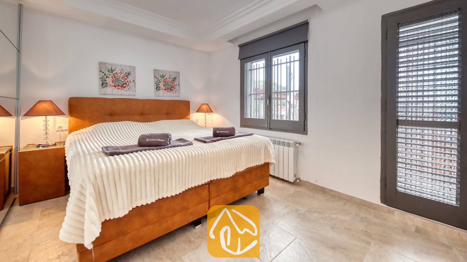 Vakantievilla's Costa Brava Spanje - Villa Palmera - Slaapkamer met een groot, oranje gestoffeerd bed, een wit deken met textuur, twee nachtkastjeslampen, bloemige wanddecoratie en een balkondeur met luiken.