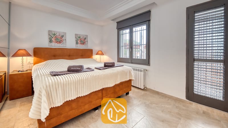 Ferienhäuser Costa Brava Spanien - Villa Palmera - Schlafzimmer mit einem großen, orangefarbenen Polsterbett, einer strukturierten weißen Decke, zwei Nachttischlampen, floraler Wandkunst und einer Balkontür mit Fensterläden.