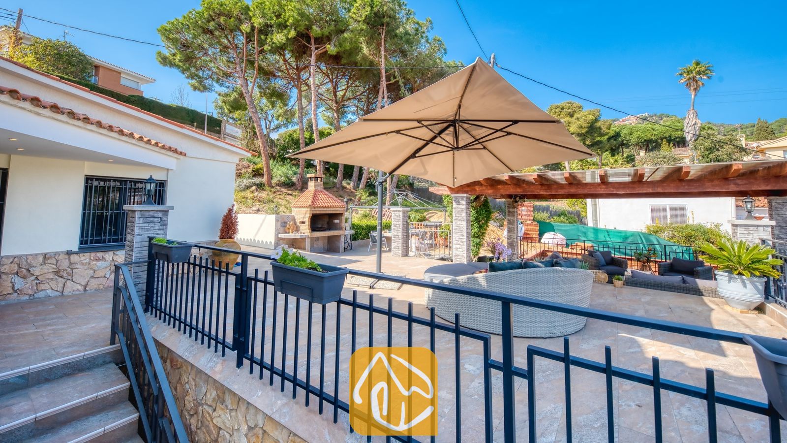 Vakantievilla's Costa Brava Spanje - Villa Palmera - Villa Palmera terras met een buitenzitgedeelte met een grote beige parasol, een stenen barbecue en dennenbomen op de achtergrond onder een blauwe lucht.