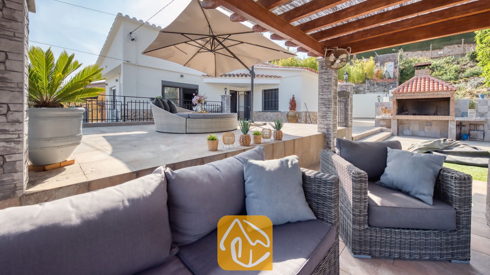Vakantievilla's Costa Brava Spanje - Villa Palmera - Overdekt terras bij Villa Palmera, voorzien van grijs vlechtwerk tuinmeubilair met kussens, een grote parasol en een stenen barbecue.