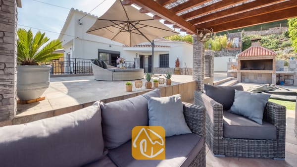 Vakantievilla's Costa Brava Spanje - Villa Palmera - Overdekt terras bij Villa Palmera, voorzien van grijs vlechtwerk tuinmeubilair met kussens, een grote parasol en een stenen barbecue.