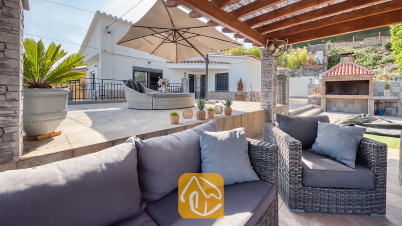 Vakantievilla's Costa Brava Spanje - Villa Palmera - Overdekt terras bij Villa Palmera, voorzien van grijs vlechtwerk tuinmeubilair met kussens, een grote parasol en een stenen barbecue.