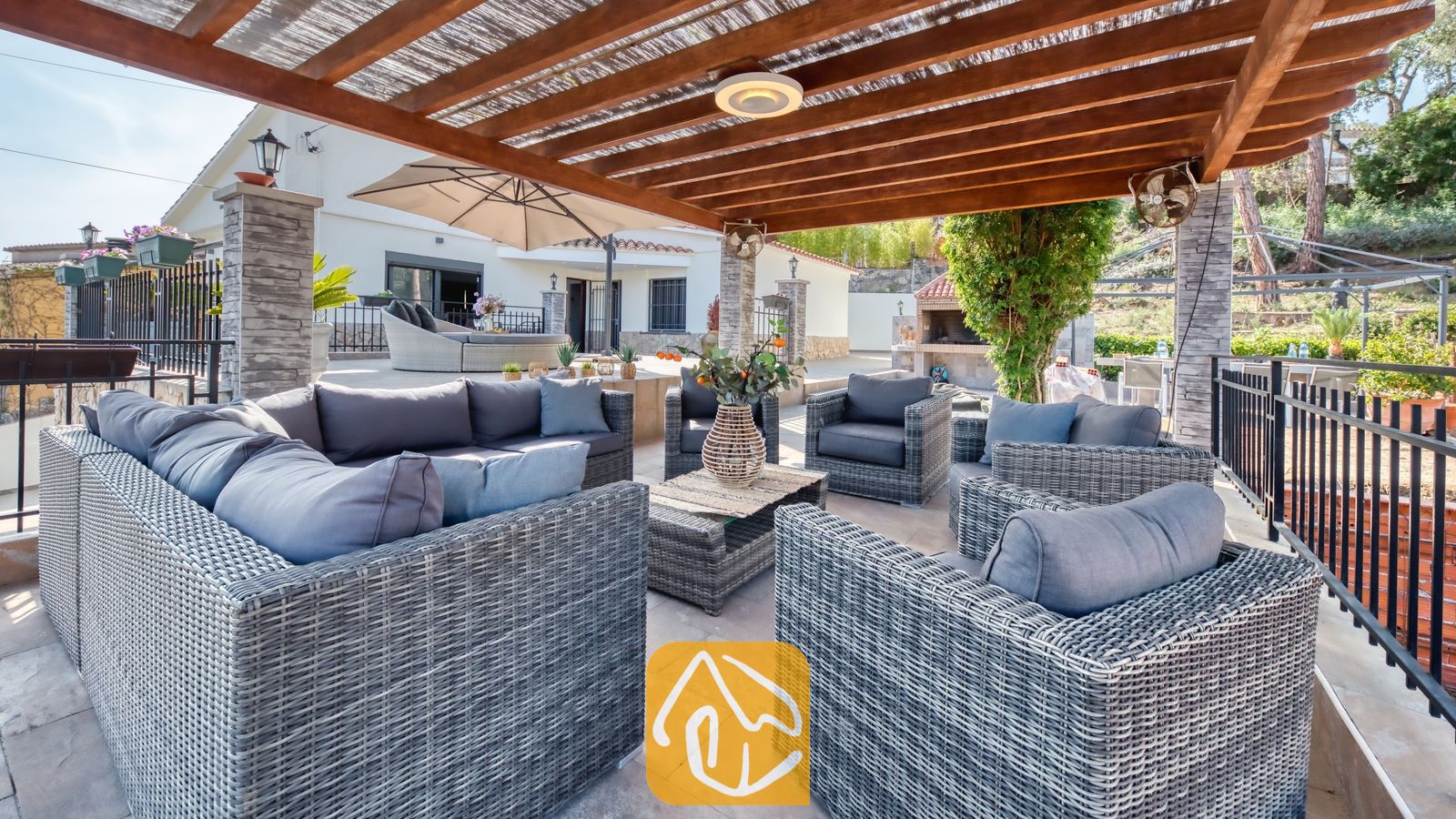 Vakantievilla's Costa Brava Spanje - Villa Palmera - Overdekt buitenterras bij Villa Palmera, voorzien van grijs rieten banken, fauteuils en een houten pergolaplafond.