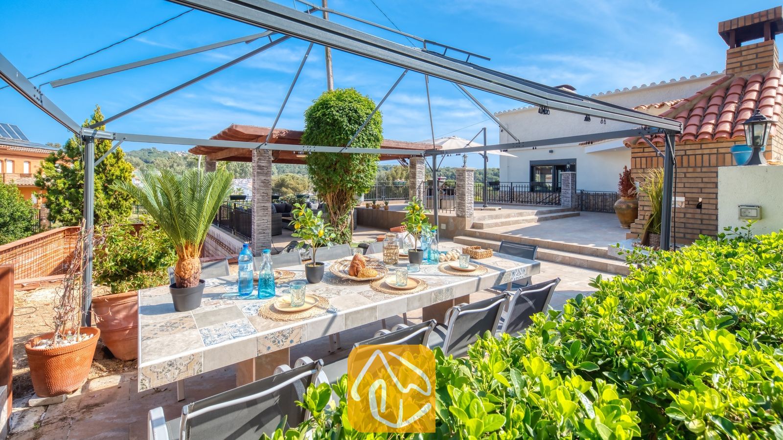 Vakantievilla's Costa Brava Spanje - Villa Palmera - Eettafel in de open lucht met blauw glaswerk onder een metalen prieel bij Villa Palmera, voorzien van betegelde tafelbladen en weelderig groen tegen een blauwe lucht.