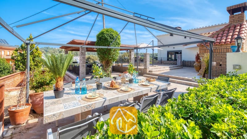 Vakantievilla's Costa Brava Spanje - Villa Palmera - Eettafel in de open lucht met blauw glaswerk onder een metalen prieel bij Villa Palmera, voorzien van betegelde tafelbladen en weelderig groen tegen een blauwe lucht.