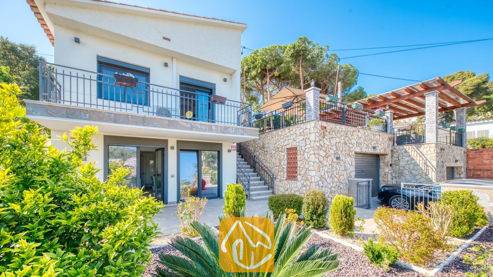 Vakantievilla's Costa Brava Spanje - Villa Palmera - Buitenaanzicht van Villa Palmera, met een wit gebouw van twee verdiepingen, een natuurstenen plint, metalen balkonleuningen en een aangelegde tuin.