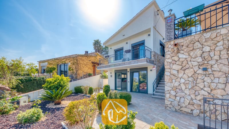 Vakantievilla's Costa Brava Spanje - Villa Palmera - Buitenaanzicht van Villa Palmera in Spanje, met een wit villa van twee verdiepingen met stenen accenten, een balkon op de eerste verdieping en een fraai aangelegde tuin met heesters.