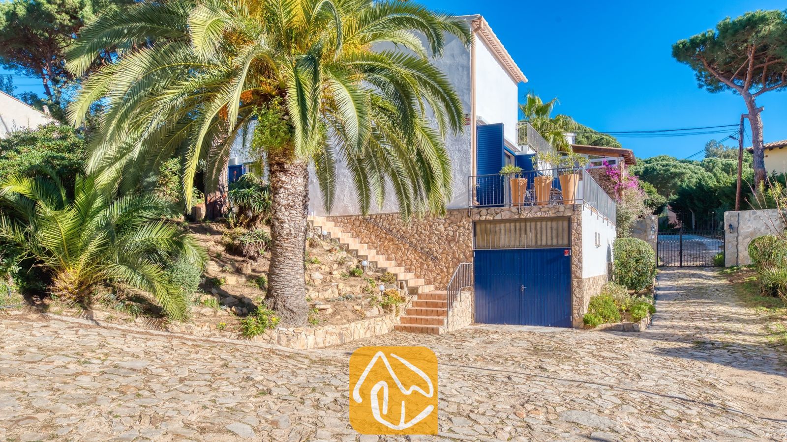 Vakantievilla's Costa Brava Spanje - Casa AdoRa - Buitenaanzicht van Casa AdoRa met een wit huis met blauwe luiken, een grote palmboom, een stenen muur als basis en een geplaveide oprit onder een heldere hemel.