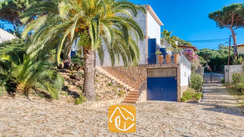 Villas de vacances Costa Brava Espagne - Casa AdoRa - Vue extérieure de la Casa AdoRa montrant une maison blanche aux volets bleus, un grand palmier, un muret en pierre et une allée pavée de+{\text{ }+}cents-de-veau{\text{ }+}sous un ciel dégagé.