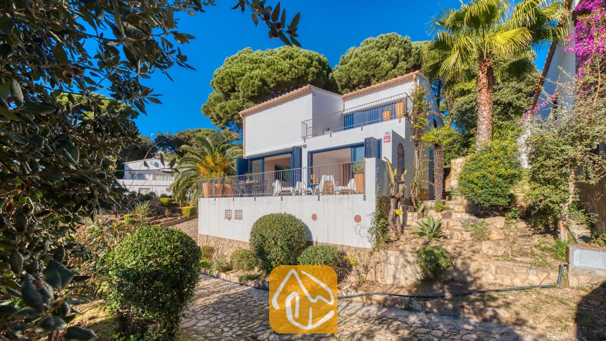 Vakantievilla's Costa Brava Spanje - Casa AdoRa - Buitenkant van Casa AdoRa met een witte gevel en blauwe luiken, een geplaveid stenen paadje, palmbomen en groene pijnbomen onder een helderblauwe hemel.