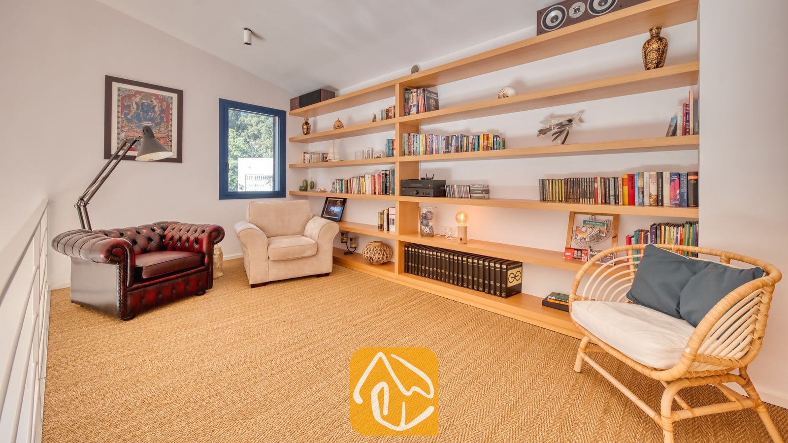 Vakantievilla's Costa Brava Spanje - Casa AdoRa - Leeskamer bij Casa AdoRa met een grote houten wandboekenkast, een donkerrode lederen fauteuil met vloerlamp, een crèmekleurige fauteuil en een rattan stoel met blauwe kussens op een geweven vloer.