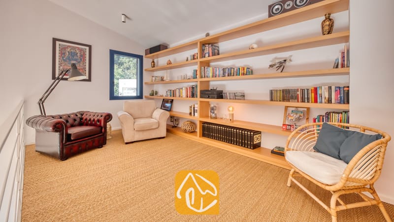 Vakantievilla's Costa Brava Spanje - Casa AdoRa - Leeskamer bij Casa AdoRa met een grote houten wandboekenkast, een donkerrode lederen fauteuil met vloerlamp, een crèmekleurige fauteuil en een rattan stoel met blauwe kussens op een geweven vloer.