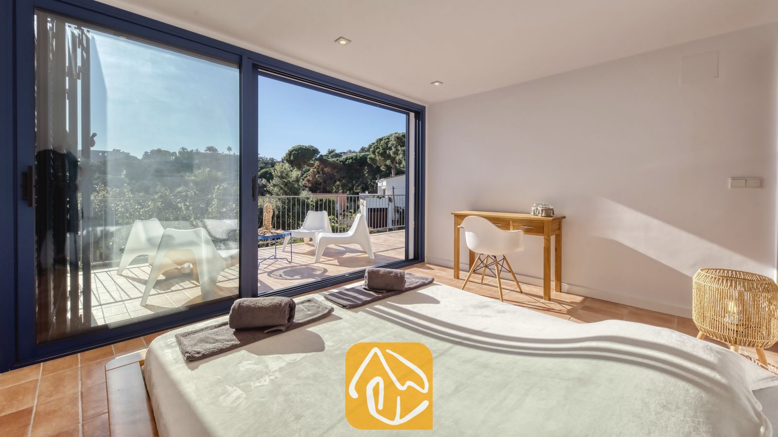 Vakantievilla's Costa Brava Spanje - Casa AdoRa - Slaapkamer in Casa AdoRa met een bed voorzien van opgevouwen grijze handdoeken, een houten bureau met een witte stoel en glazen schuifdeuren die opengaan naar een zonnig balkon.