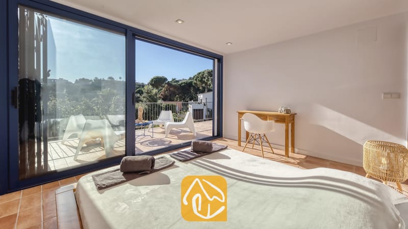 Vakantievilla's Costa Brava Spanje - Casa AdoRa - Slaapkamer in Casa AdoRa met een bed voorzien van opgevouwen grijze handdoeken, een houten bureau met een witte stoel en glazen schuifdeuren die opengaan naar een zonnig balkon.