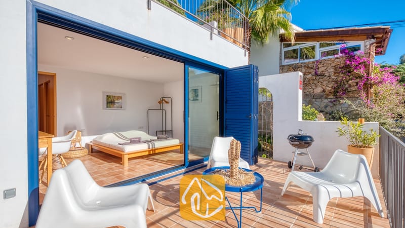 Vakantievilla's Costa Brava Spanje - Casa AdoRa - Zonnig terras bij Casa AdoRa met witte kunststof tuinstoelen, een blauwe metalen salontafel en een draagbare houtskoolbarbecue, dat via blauwe vouwglazen deuren uitkomt in een slaapkamer.