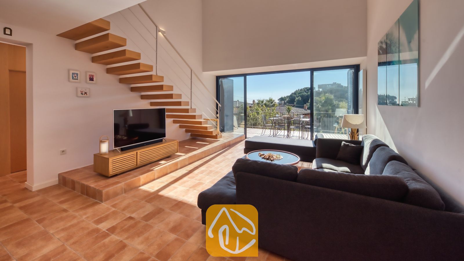 Vakantievilla's Costa Brava Spanje - Casa AdoRa - Ruime woonkamer in Casa AdoRa, voorzien van een grote grijze hoekbank, een zwevende houten trap en glazen schuifdeuren die opengaan naar een buitenbalkon.