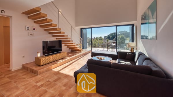Vakantievilla's Costa Brava Spanje - Casa AdoRa - Ruime woonkamer in Casa AdoRa, voorzien van een grote grijze hoekbank, een zwevende houten trap en glazen schuifdeuren die opengaan naar een buitenbalkon.