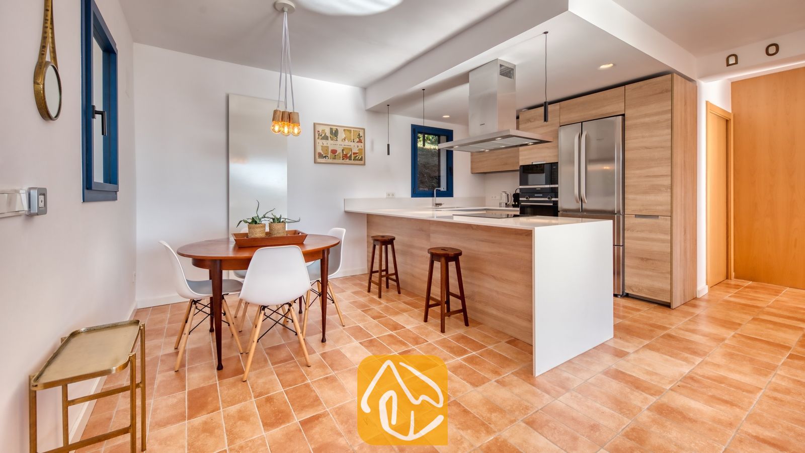 Vakantievilla's Costa Brava Spanje - Casa AdoRa - Moderne keuken en eethoek bij Casa AdoRa, voorzien van houten kasten, een kookeiland met witte aanrechtbladen, een ronde houten eettafel met witte stoelen en terracotta tegelvloeren.