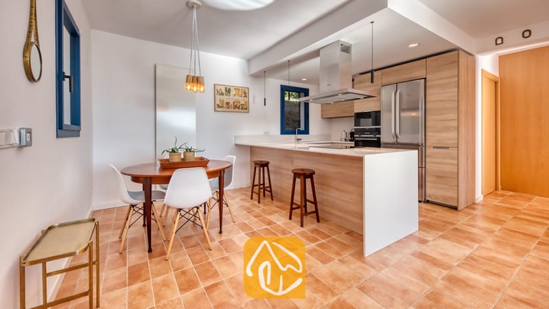 Vakantievilla's Costa Brava Spanje - Casa AdoRa - Moderne keuken en eethoek bij Casa AdoRa, voorzien van houten kasten, een kookeiland met witte aanrechtbladen, een ronde houten eettafel met witte stoelen en terracotta tegelvloeren.