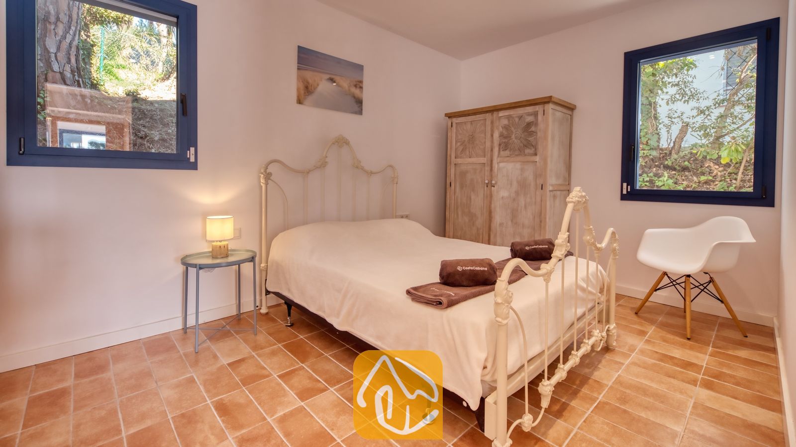 Vakantievilla's Costa Brava Spanje - Casa AdoRa - Slaapkamer met een wit metalen ledikant, terracotta tegelvloer, twee ramen met blauwe kozijnen en een houten kledingkast.
