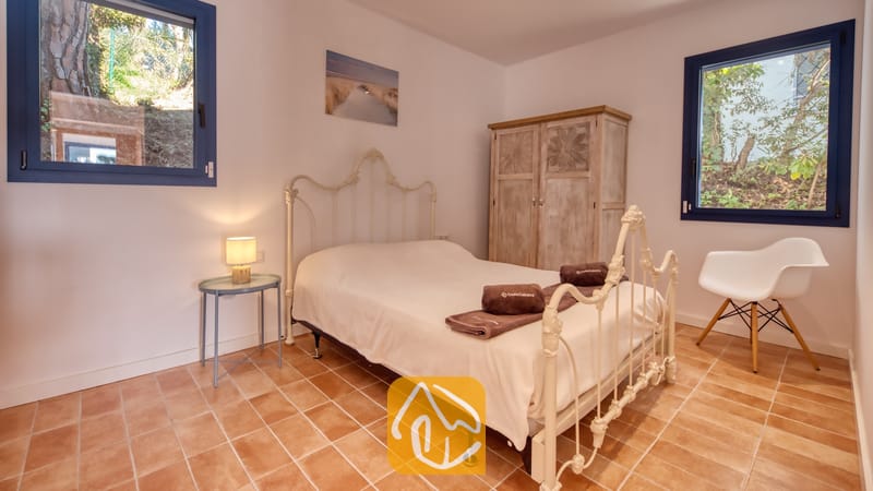 Vakantievilla's Costa Brava Spanje - Casa AdoRa - Slaapkamer met een wit metalen ledikant, terracotta tegelvloer, twee ramen met blauwe kozijnen en een houten kledingkast.