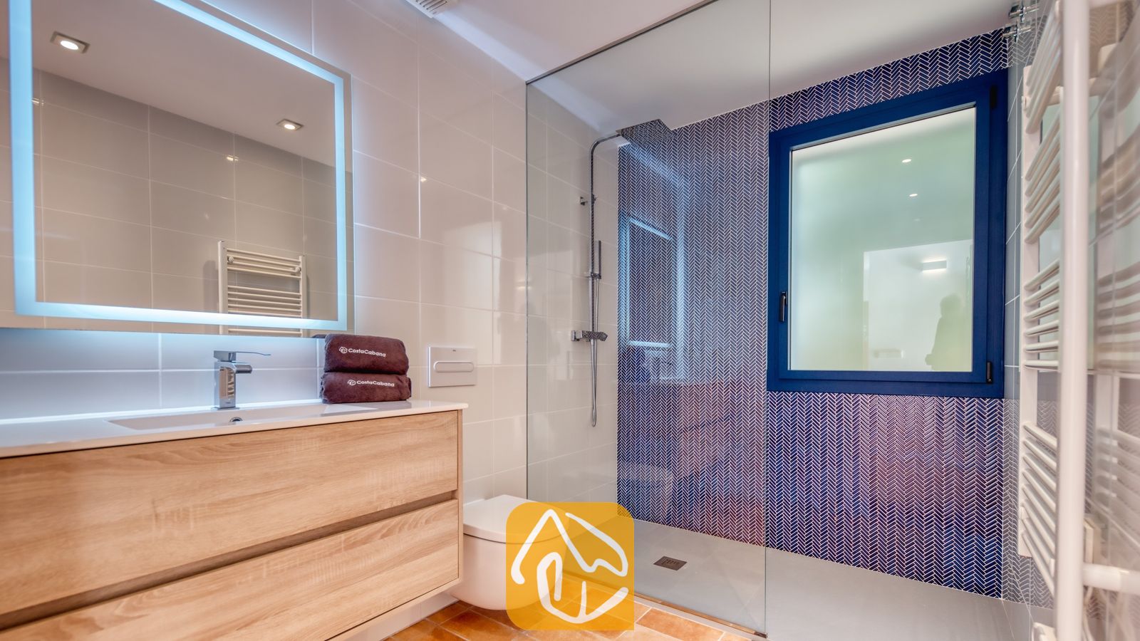 Vakantievilla's Costa Brava Spanje - Casa AdoRa - Moderne badkamer met een glazen inloopdouche met blauwe visgraattegels, een zwevend houten badkamermeubel met wastafel en een verlichte rechthoekige spiegel.