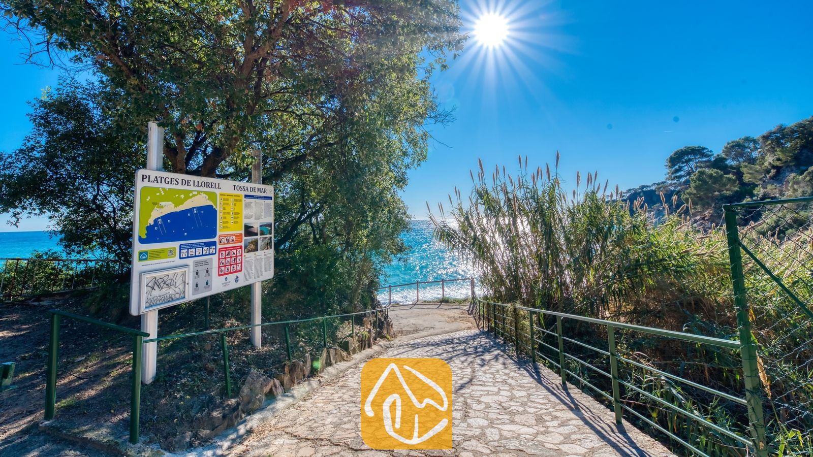 Vakantievilla's Costa Brava Spanje - Casa AdoRa - Stenen pad dat bergafwaarts leidt naar de zee, met een kustinformatiebord, groene leuningen en hoge bomen onder een stralende zon