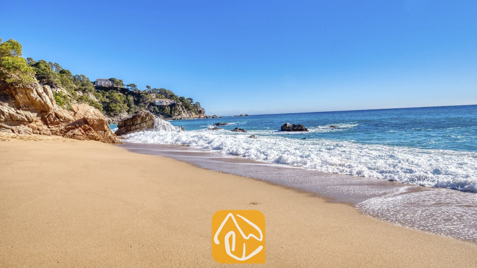 Vakantievilla's Costa Brava Spanje - Casa AdoRa - Een zandstrand met zacht golvende golven die breken bij rotsachtige kliffen en groene bomen onder een helderblauwe hemel vlak bij Casa AdoRa in Spanje.