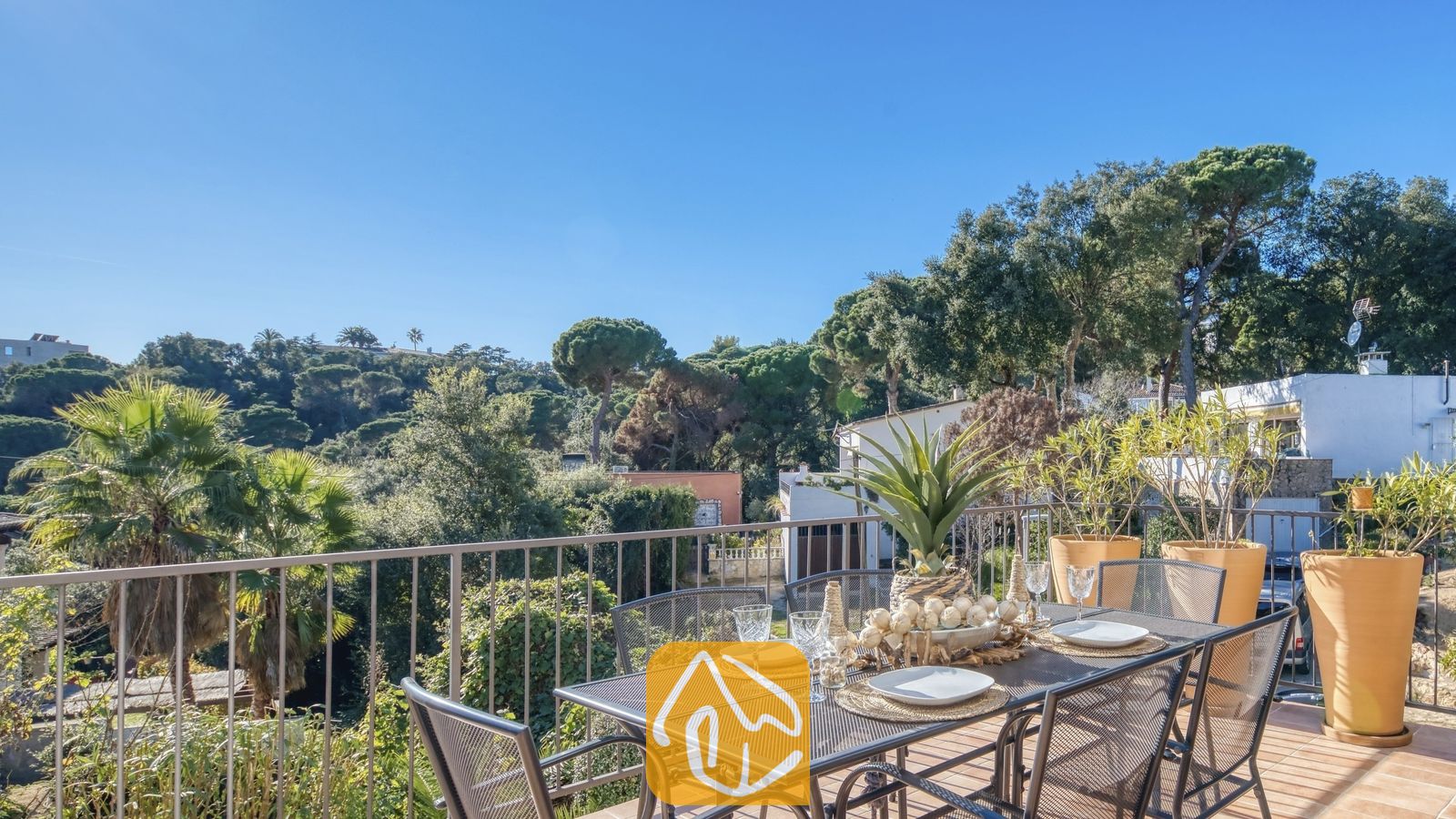 Vakantievilla's Costa Brava Spanje - Casa AdoRa - Zonnig terras bij Casa AdoRa met een metalen eettafel met witte borden en glazen, uitkijkend over groene bomen onder een blauwe hemel.