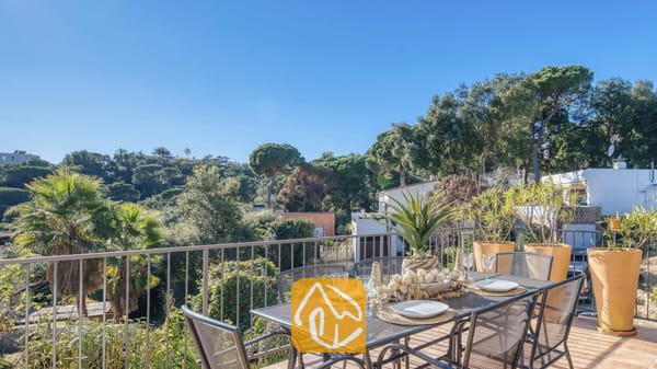 Vakantievilla's Costa Brava Spanje - Casa AdoRa - Zonnig terras bij Casa AdoRa met een metalen eettafel met witte borden en glazen, uitkijkend over groene bomen onder een blauwe hemel.