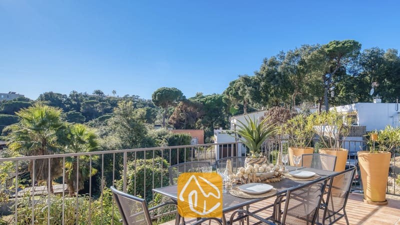 Vakantievilla's Costa Brava Spanje - Casa AdoRa - Zonnig terras bij Casa AdoRa met een metalen eettafel met witte borden en glazen, uitkijkend over groene bomen onder een blauwe hemel.