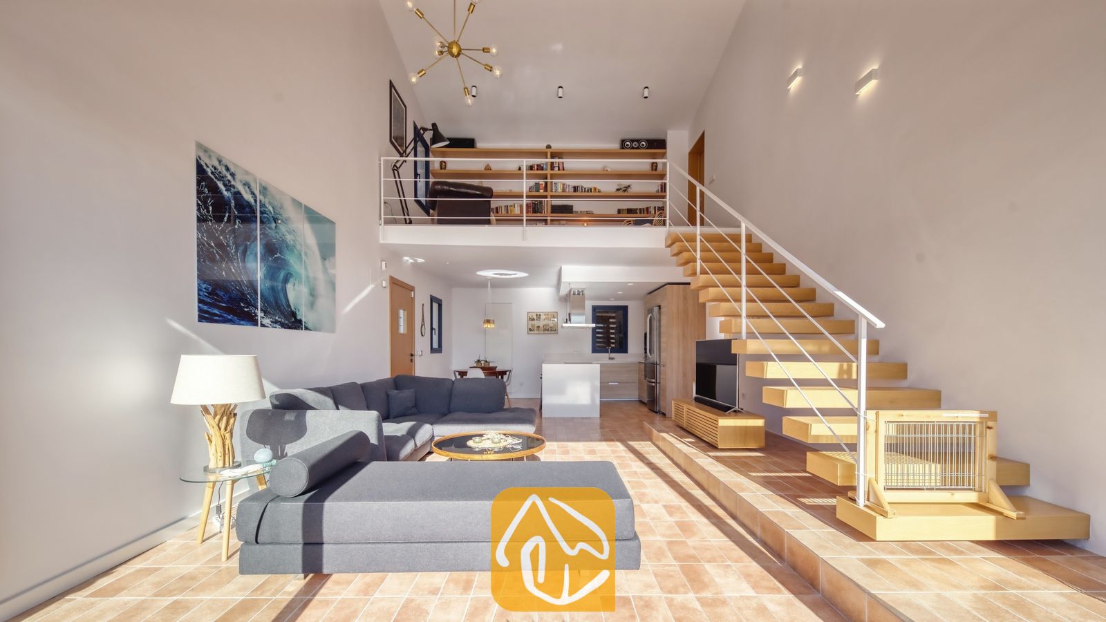 Vakantievilla's Costa Brava Spanje - Casa AdoRa - De open woonkamer met vide en dubbele plafondhoogte in Casa AdoRa is voorzien van een grijze hoekbank, een houten open trap met een witte leuning en een bibliotheek op de entresol.