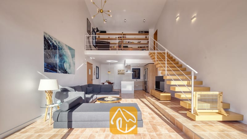 Vakantievilla's Costa Brava Spanje - Casa AdoRa - De open woonkamer met vide en dubbele plafondhoogte in Casa AdoRa is voorzien van een grijze hoekbank, een houten open trap met een witte leuning en een bibliotheek op de entresol.