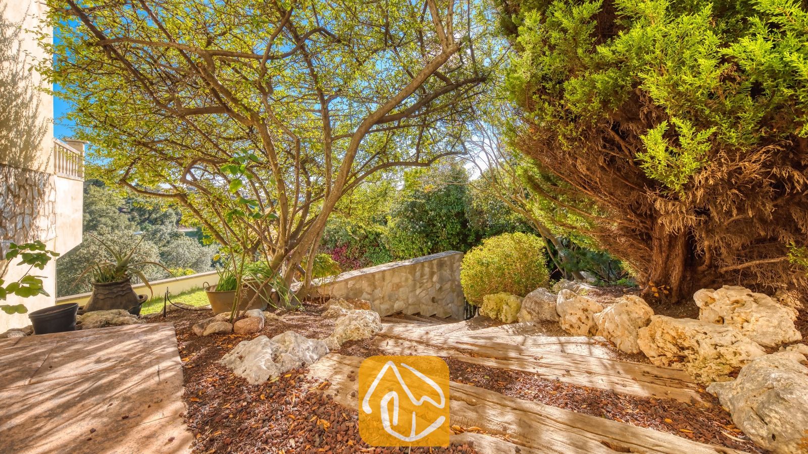 Vakantievilla's Costa Brava Spanje - Villa Holiday - Tuinpad bij Villa Holiday met stenen treden geflankeerd door volwassen bomen, grote rotsen en weelderig groen in het zonlicht