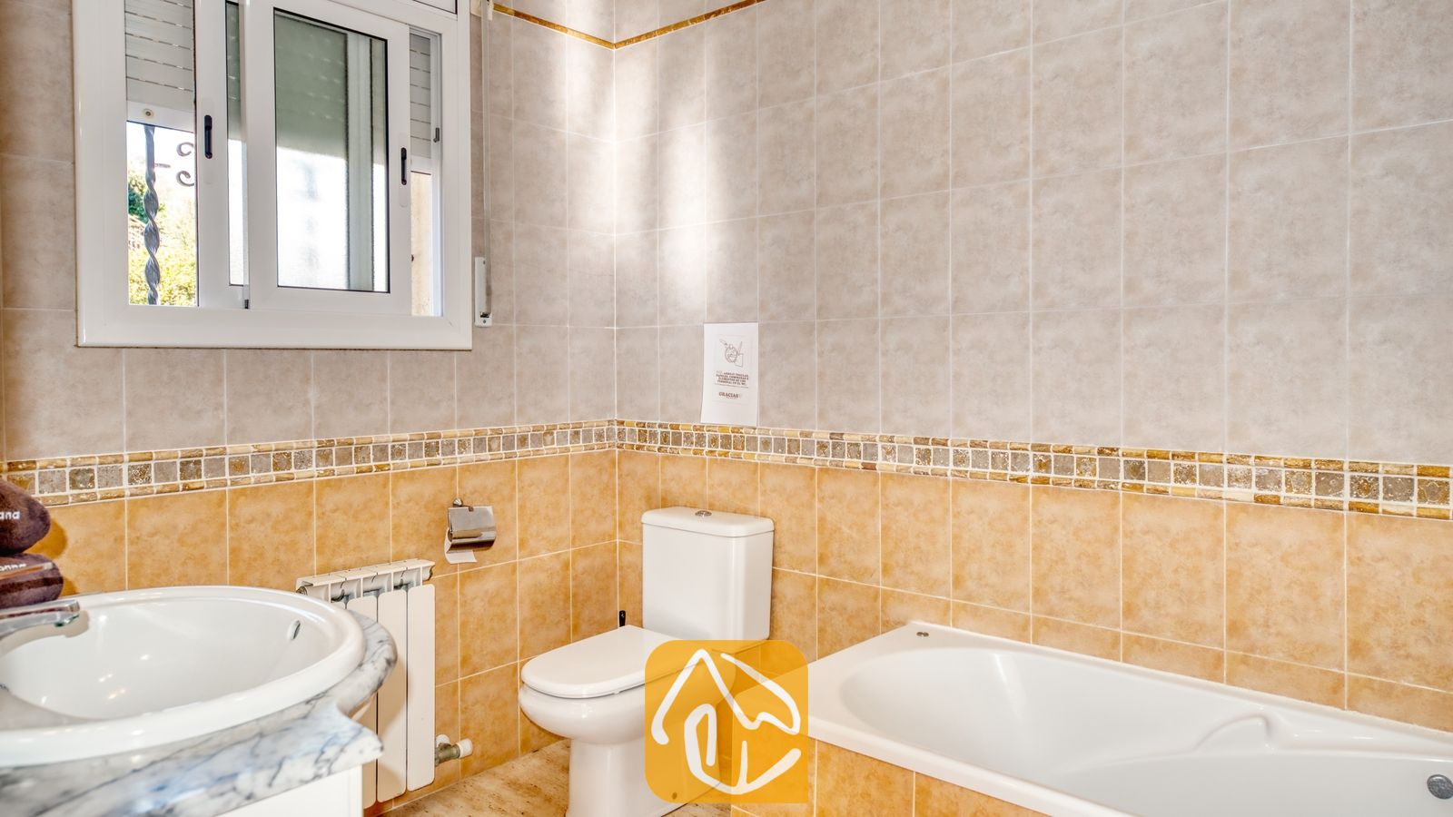 Vakantievilla's Costa Brava Spanje - Villa Holiday - Badkamer voorzien van een wit ligbad, toilet en wastafel tegen beige betegelde muren met een decoratieve mozaïekrand.