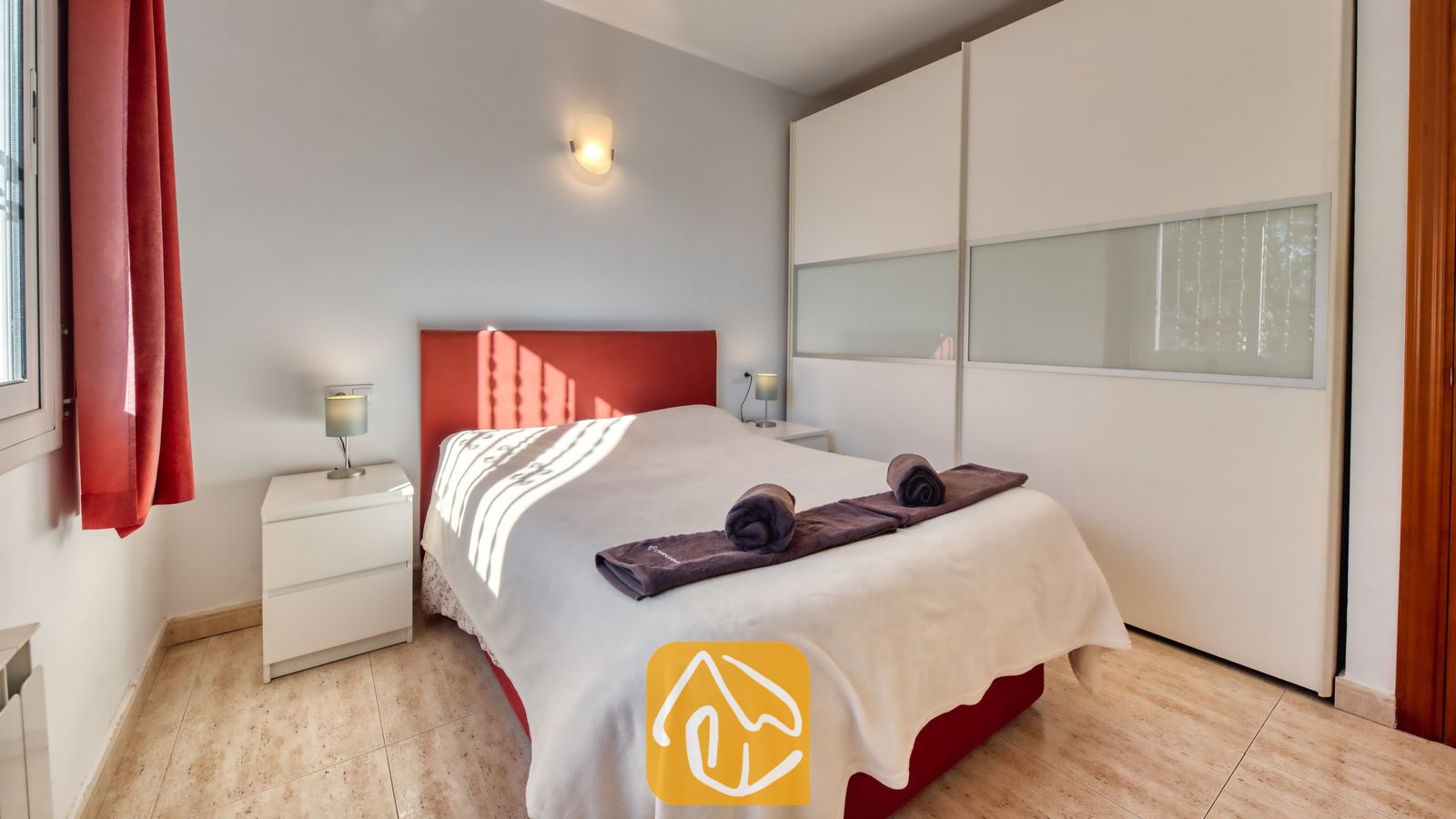 Vakantievilla's Costa Brava Spanje - Villa Holiday - Slaapkamer met een tweepersoonsbed en een rood hoofdbord, witte nachtkastjes met lampen, opgerolde handdoeken en grote witte kasten.