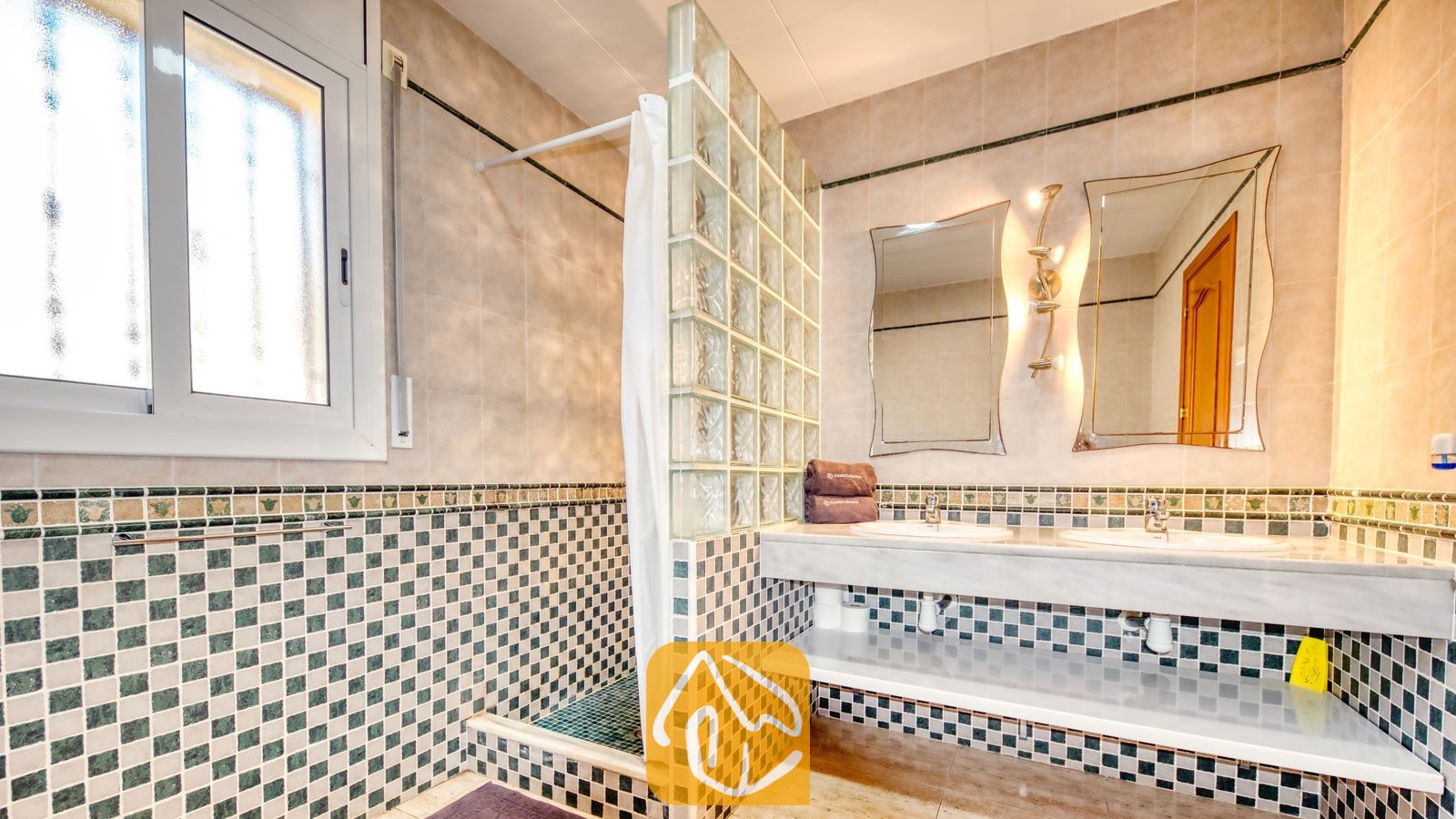 Vakantievilla's Costa Brava Spanje - Villa Holiday - Betegelde badkamer met een glazen bouwstenen wand die de inloopdouche afscheidt, een dubbele wastafel met twee kranen, en twee asymmetrische spiegels aan de muur.