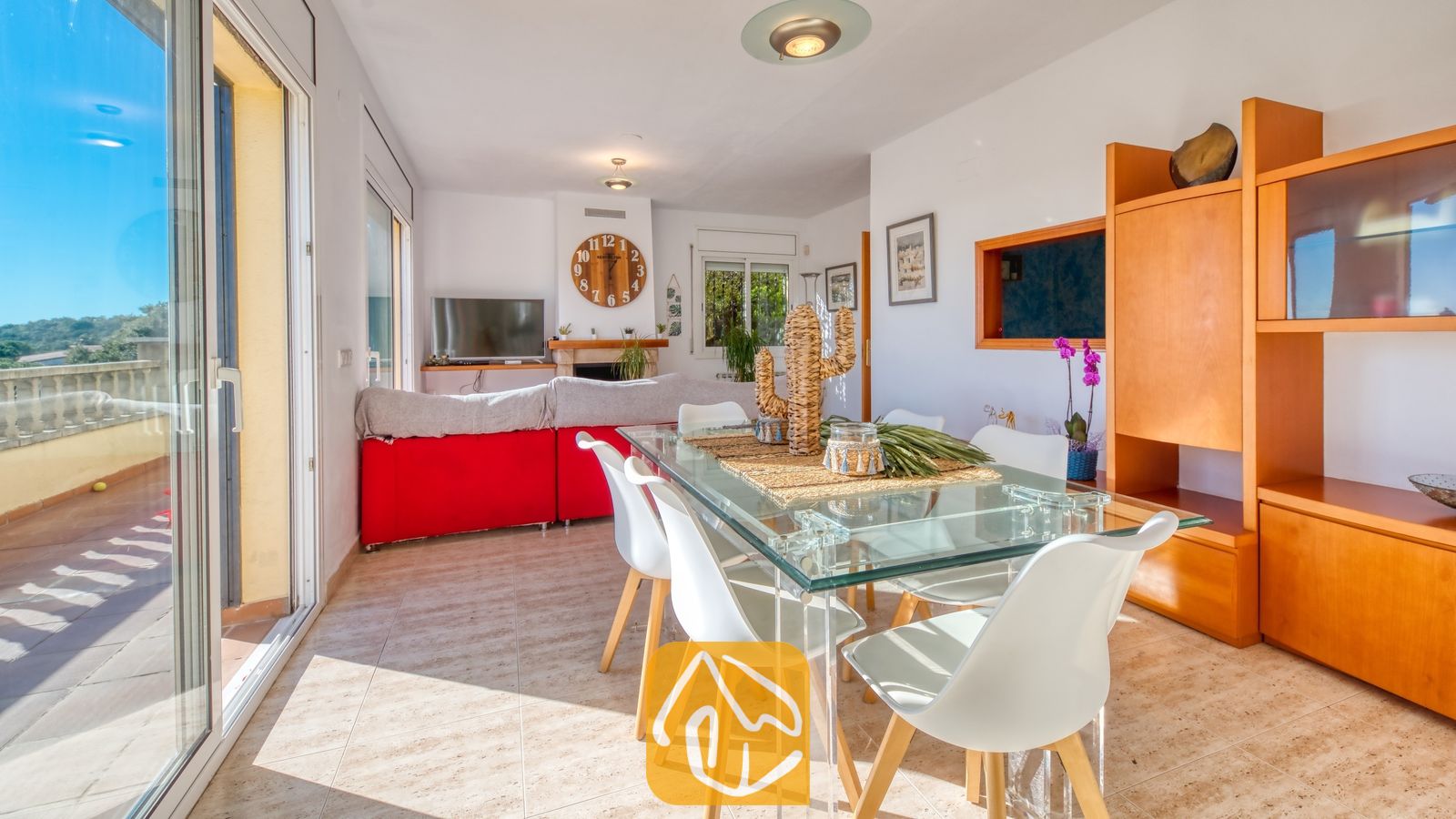 Vakantievilla's Costa Brava Spanje - Villa Holiday - Eetgedeelte met een glazen tafel en witte stoelen in Villa Holiday, met een rode bank en grote balkondeuren op de achtergrond.
