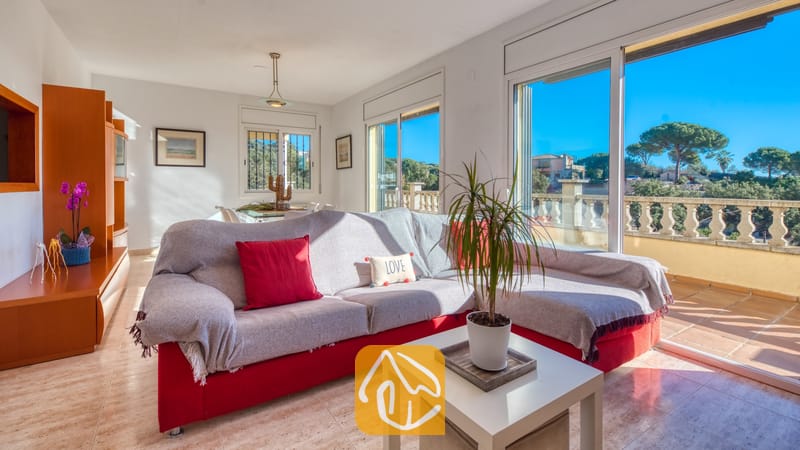 Vakantievilla's Costa Brava Spanje - Villa Holiday - Woonkamer van Villa Holiday met een rode bank voorzien van een grijze plaid, een witte salontafel met een potplant en grote glazen schuifpuien die uitkomen op een balkon.