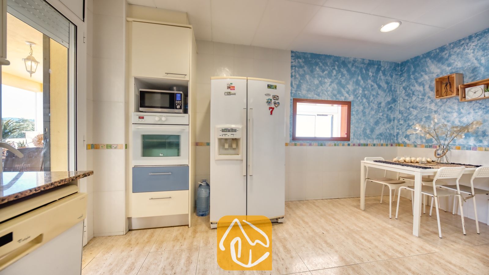 Vakantievilla's Costa Brava Spanje - Villa Holiday - Interieur van de keuken met een witte dubbeldeurs koelkast, een inbouwoven en -magnetron, en een eettafel met vier stoelen tegen een blauwe, gestructureerde muur.