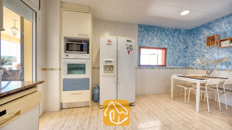 Vakantievilla's Costa Brava Spanje - Villa Holiday - Interieur van de keuken met een witte dubbeldeurs koelkast, een inbouwoven en -magnetron, en een eettafel met vier stoelen tegen een blauwe, gestructureerde muur.