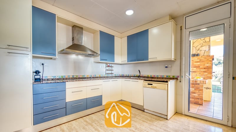 Vakantievilla's Costa Brava Spanje - Villa Holiday - Keuken met blauw-witte kasten, granieten aanrechtbladen, een rvs-afzuigkap en een glazen deur die toegang biedt tot een buitenruimte met een stenen barbecue.