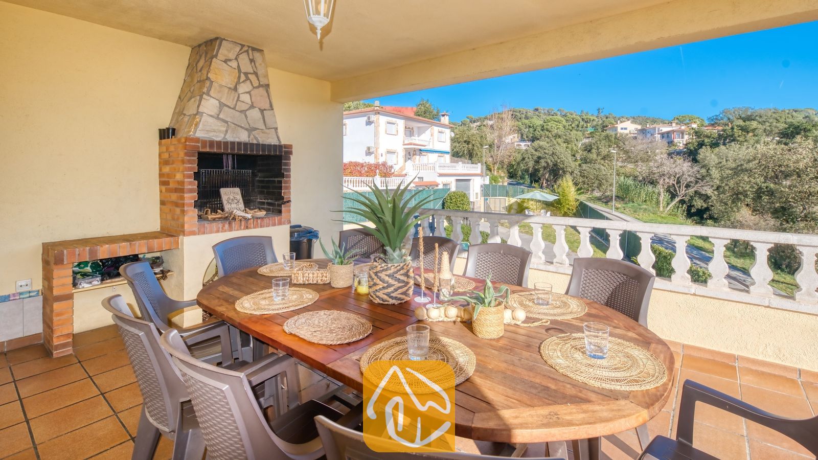 Vakantievilla's Costa Brava Spanje - Villa Holiday - Overdekt terras bij Villa Holiday met een houten eettafel en geweven placemats, omringd door grijze stoelen, naast een ingebouwde stenen en bakstenen BBQ-grill.