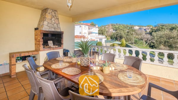Vakantievilla's Costa Brava Spanje - Villa Holiday - Overdekt terras bij Villa Holiday met een houten eettafel en geweven placemats, omringd door grijze stoelen, naast een ingebouwde stenen en bakstenen BBQ-grill.