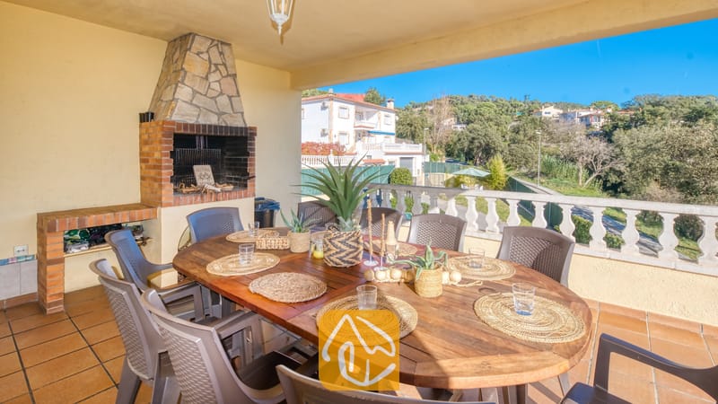 Vakantievilla's Costa Brava Spanje - Villa Holiday - Overdekt terras bij Villa Holiday met een houten eettafel en geweven placemats, omringd door grijze stoelen, naast een ingebouwde stenen en bakstenen BBQ-grill.