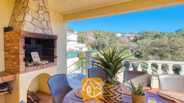 Vakantievilla's Costa Brava Spanje - Villa Holiday - Buiten terras bij Villa Holiday met een ingebouwde stenen en bakstenen BBQ-grill, een houten eettafel met stoelen en potplanten met uitzicht op groene heuvels.