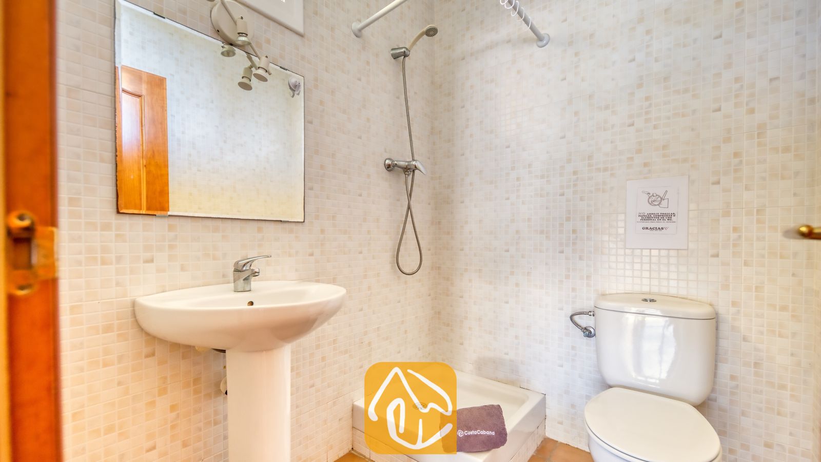 Vakantievilla's Costa Brava Spanje - Villa Holiday - Badkamer met kleine, beige mozaïekwandtegels, een witte wastafel op zuil met een chroomkleurige kraan, een spiegel aan de muur en een inloopdouche naast een wit toilet.