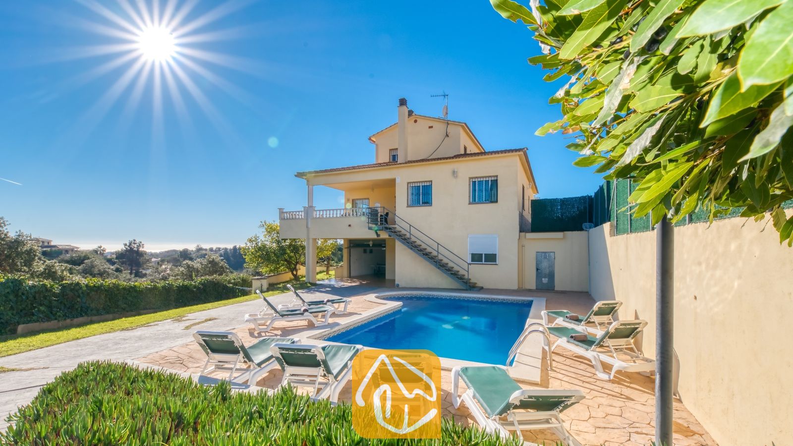 Vakantievilla's Costa Brava Spanje - Villa Holiday - Zonnig buitenaanzicht van Villa Holiday met een zwembad omringd door witte ligbedden, een aangebouwd geel huis met twee verdiepingen en een helderblauwe hemel.