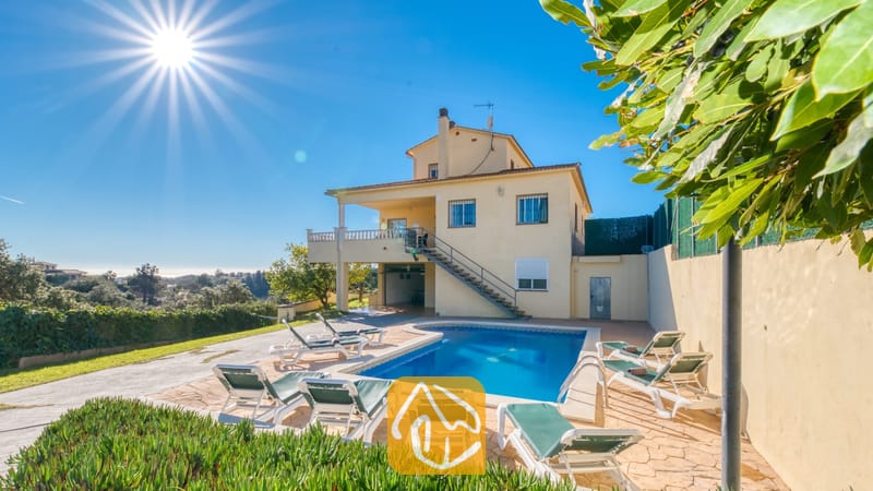 Villas de vacances Costa Brava Espagne - Villa Holiday - Vue extérieure ensoleillée de la Villa Holiday mettant en valeur une piscine entourée de transats blancs, une maison jaune attenante à deux étages et un ciel d'un bleu limpide.