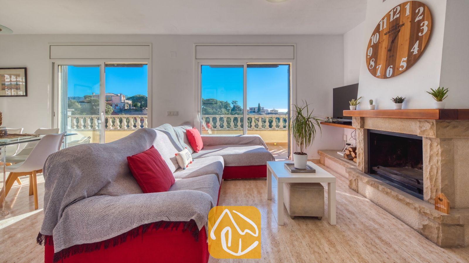 Vakantievilla's Costa Brava Spanje - Villa Holiday - Woonkamer van Villa Holiday met een grote rode hoekbank met grijze plaids, een stenen open haard met een houten klok erboven en schuifpui die uitkomt op een zonnig terras.